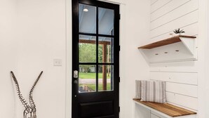 Una puerta negra con un panel de vidrio, un banco de madera con cojín y una pared blanca con líneas horizontales.