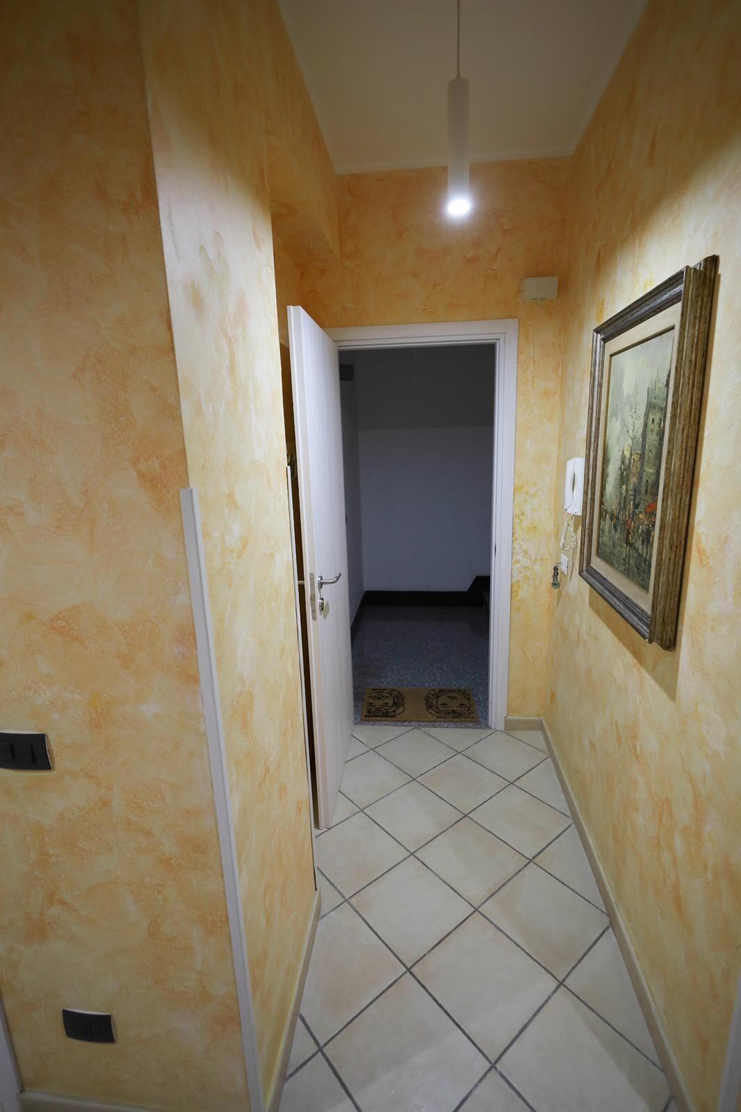Ein schmaler Korridor mit Fliesenboden, einer Tür zu einer Treppe und einem gerahmten Bild an der Wand.
