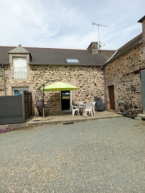 A stone building with a patio area featuring a table and chairs, a green umbrella, and a small dog. - Gîte "aux nids d'hirondelles", 8 kms from the beach. (Hénansal)