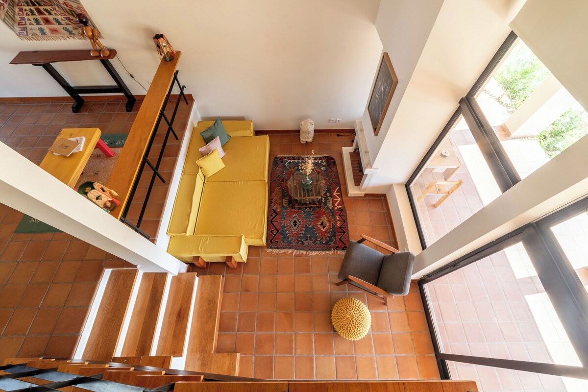 A spacious living area with a high ceiling, a wooden staircase, and a large window providing natural light.