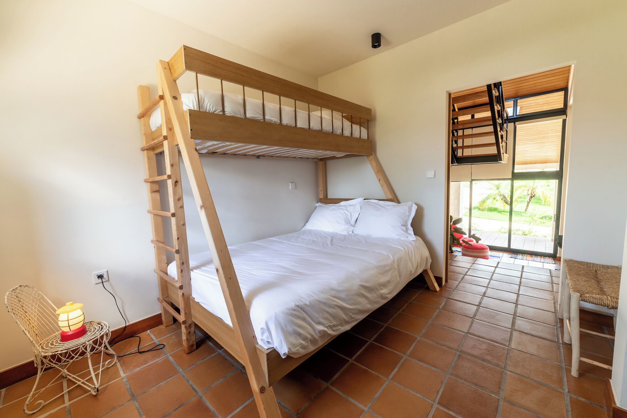A bunk bed room with a ladder, a chair with a lamp, and a view of greenery through a window.