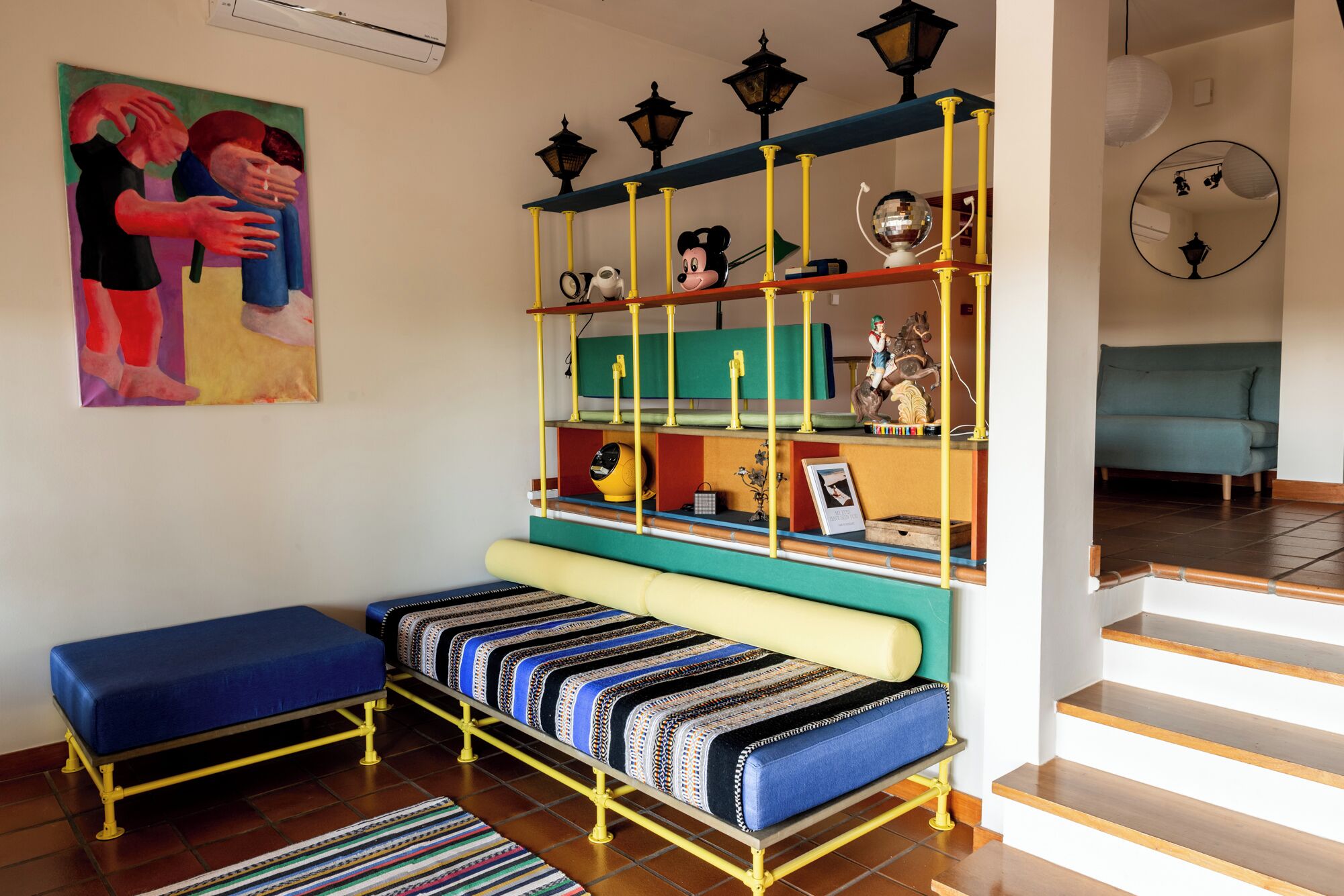 A room with a blue bench, a colorful painting, a shelf with various items, and a staircase.