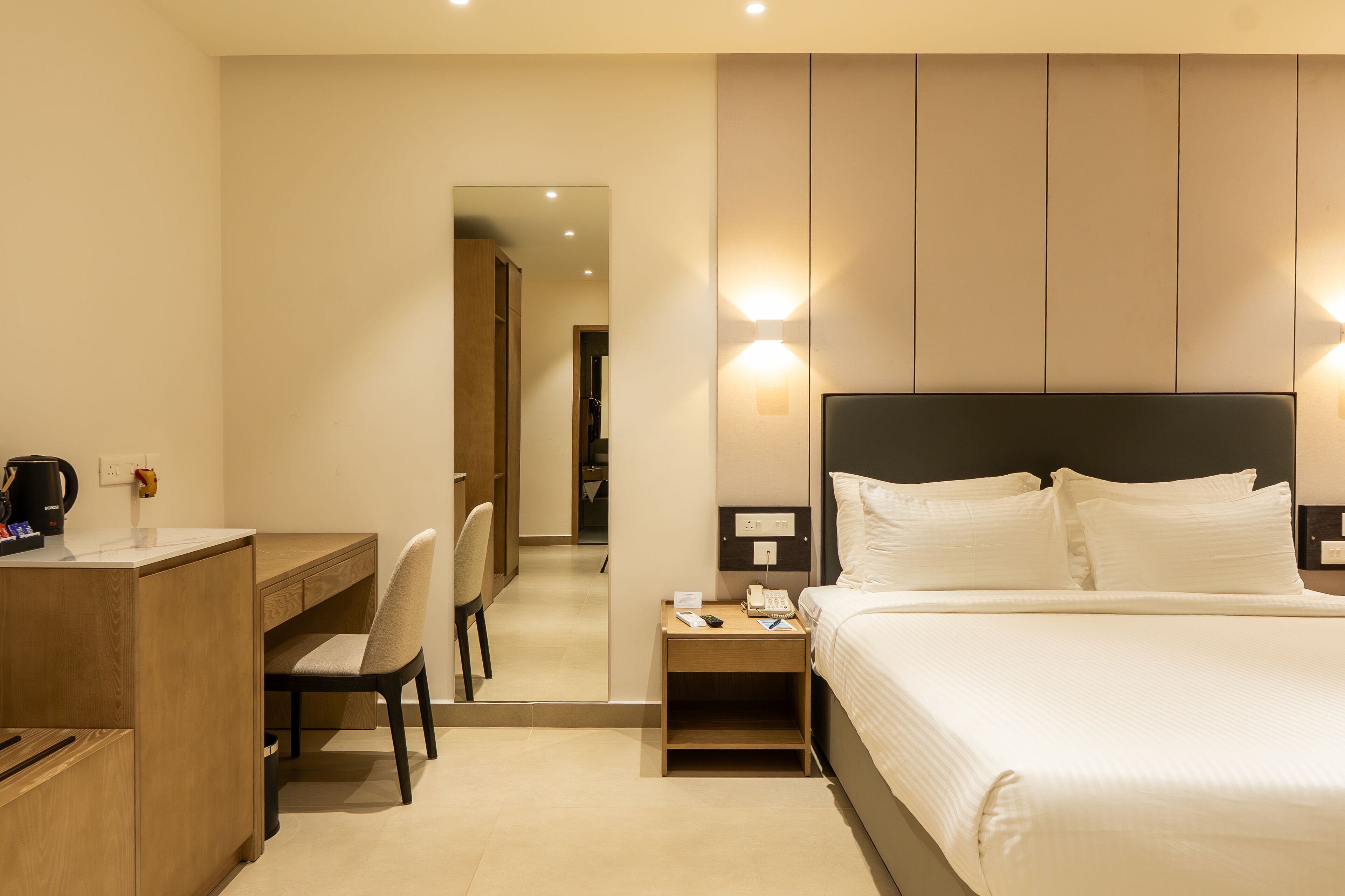 A hotel room with a bed, a desk, a chair, a nightstand, and a mirror.