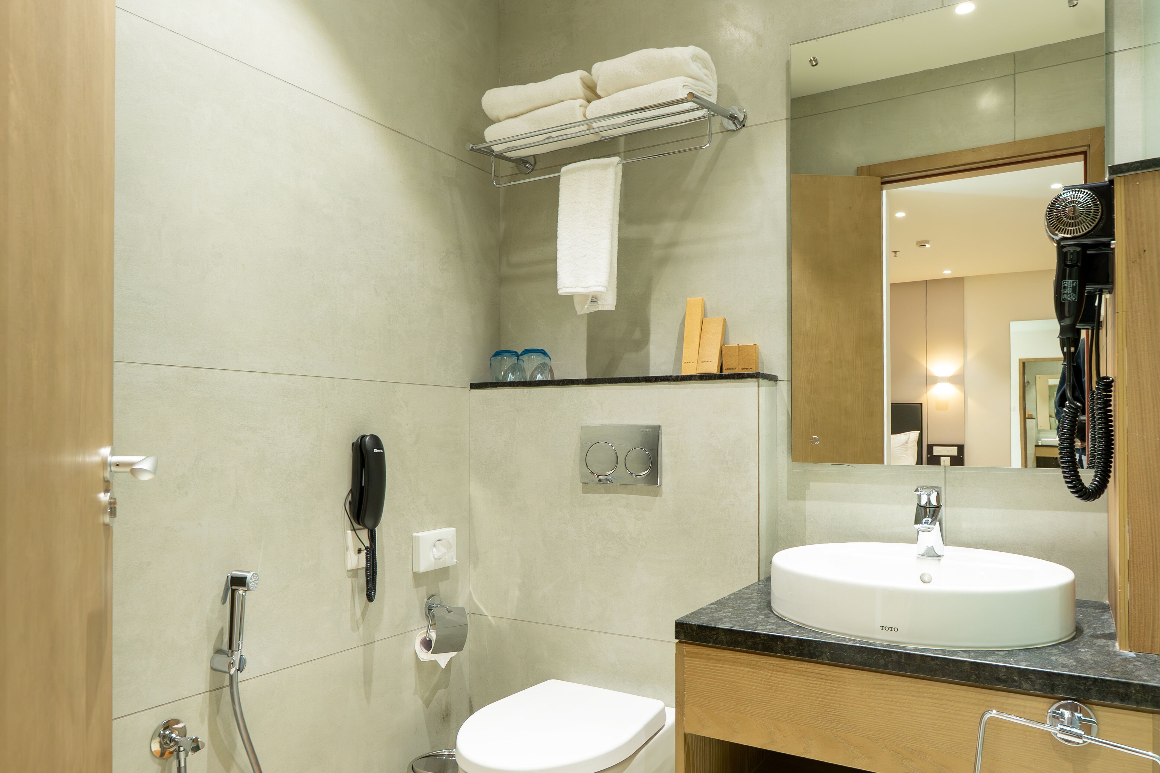 A modern bathroom with a sink, toilet, and shower. There is a towel rack with folded towels, a hairdryer, and a phone on the wall.