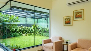 A room with a large glass wall providing a view of a well-maintained garden. There are two chairs and a small table with a plant. - Riviera Garden (Kanayannur)