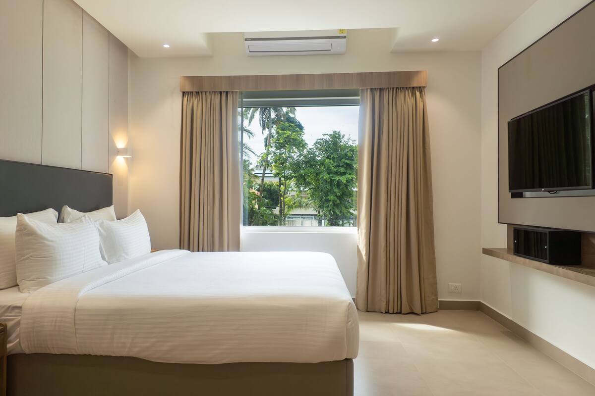 A hotel room with a large bed, a flat-screen TV mounted on the wall, and a window with curtains.