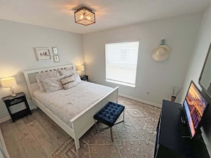 A bedroom with a bed, bedside tables, a bench, a TV, and a window with blinds. - Anastasia by the Sea 208A – Where Plans Meet Possibilities (St. Augustine)
