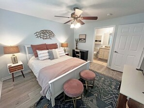 A bedroom with a bed, bedside tables, a ceiling fan, a nightstand, a desk, two stools, and a bathroom visible through an open door.