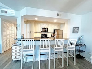 A modern kitchen with white cabinets, a central island with bar stools, and a stainless steel refrigerator. - Anastasia by the Sea 208A – Where Plans Meet Possibilities (St. Augustine)