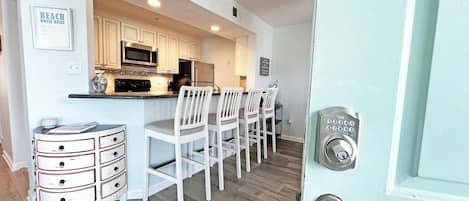 A kitchen with white cabinets, a microwave, and a refrigerator. There are bar stools and a white cabinet with drawers.