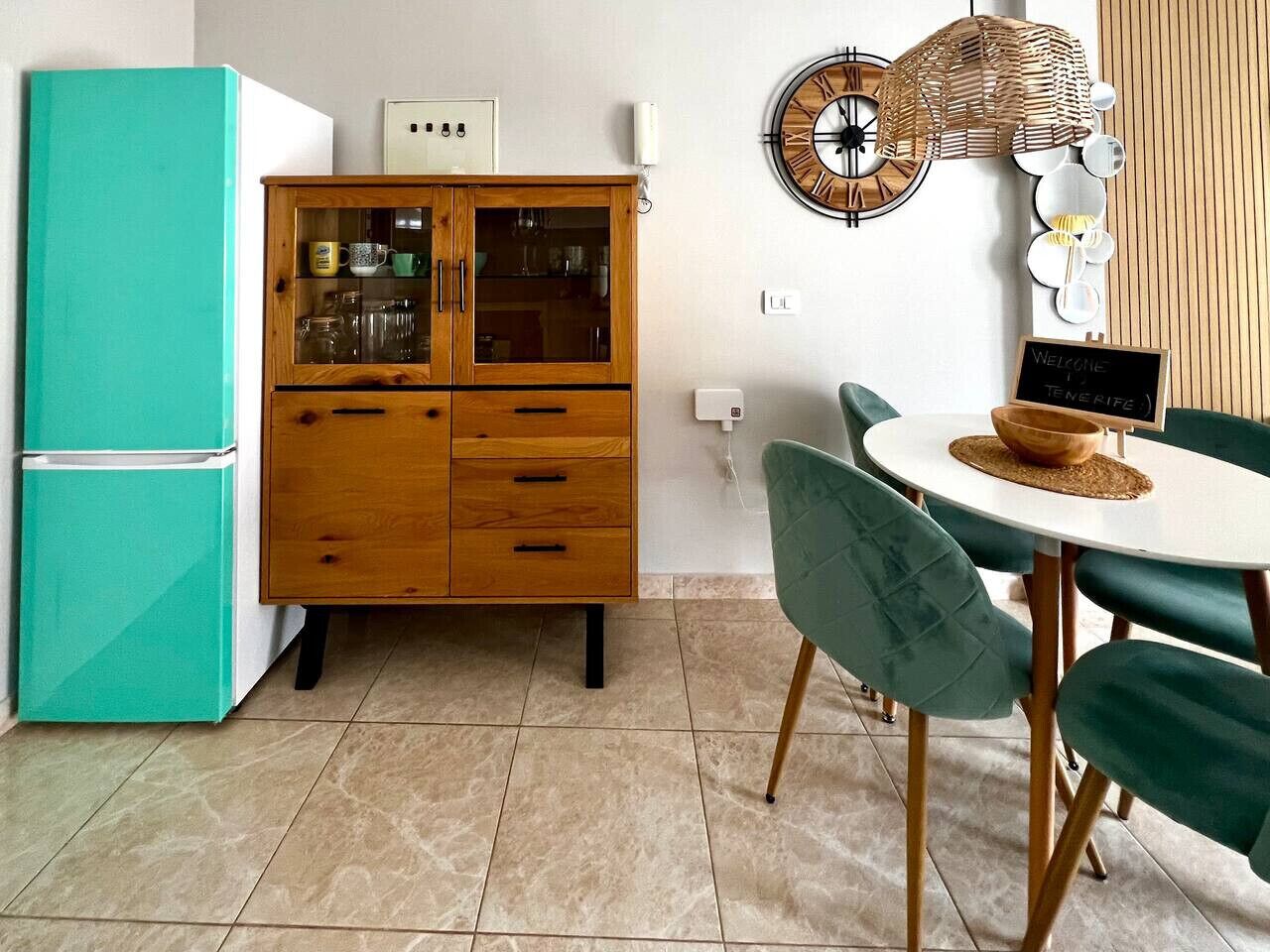 Eine Küche mit einem türkisblauen Kühlschrank, Holzschränken und einem Essbereich mit rundem Tisch und grünen Stühlen.