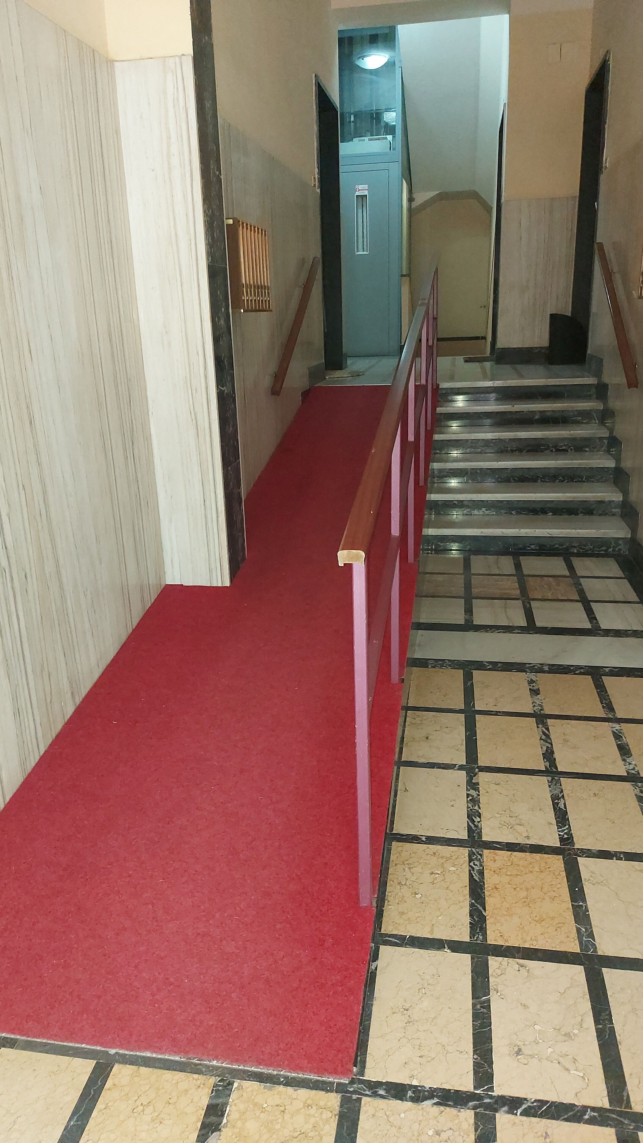 A staircase with a red carpet leading up, flanked by a wooden handrail and a tiled floor with a black and beige pattern.
