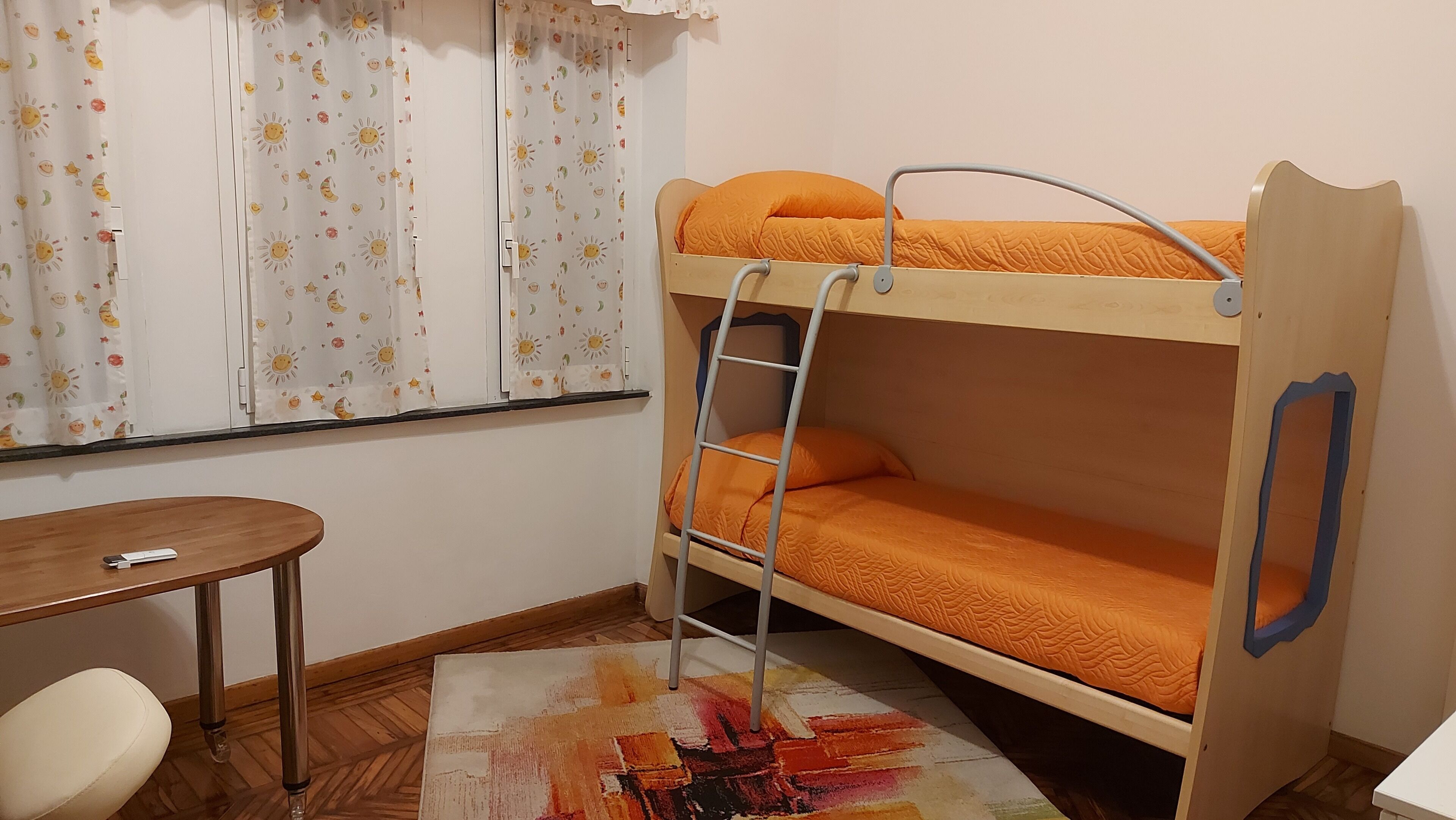 Ein Etagenbett mit orangefarbener Matratze, einer Stehleiter und einem Holztisch mit Fernbedienung.