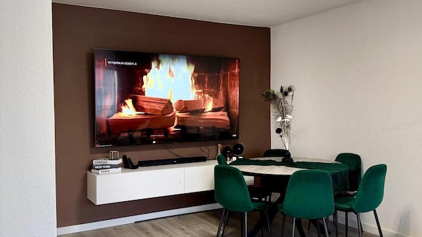 A modern living room with a flat-screen TV mounted on a wall, a dining table with green chairs, and a minimalist design.