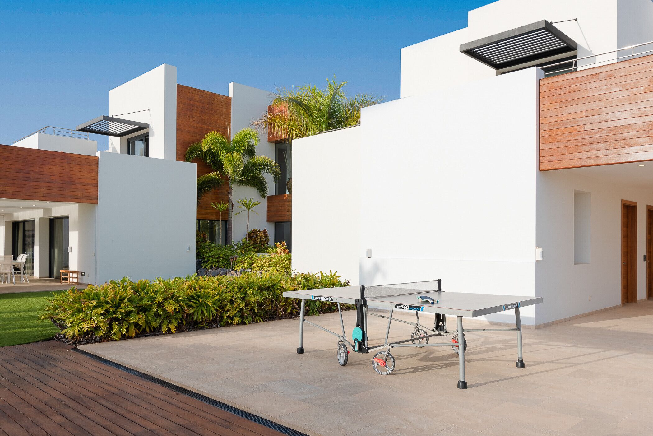 A modern outdoor area with a ping pong table, a wooden deck, and a white building with a wooden facade.
