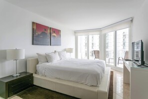 A modern bedroom with a bed, bedside lamps, a desk, and a large window with a view of buildings.