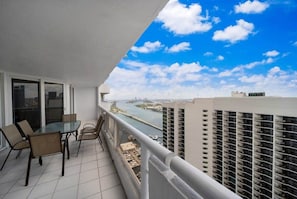 A balcony with a glass table and chairs, overlooking a river and cityscape.
