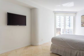 Quarto com cama, televisão fixada na parede e janela com vista para edifícios.