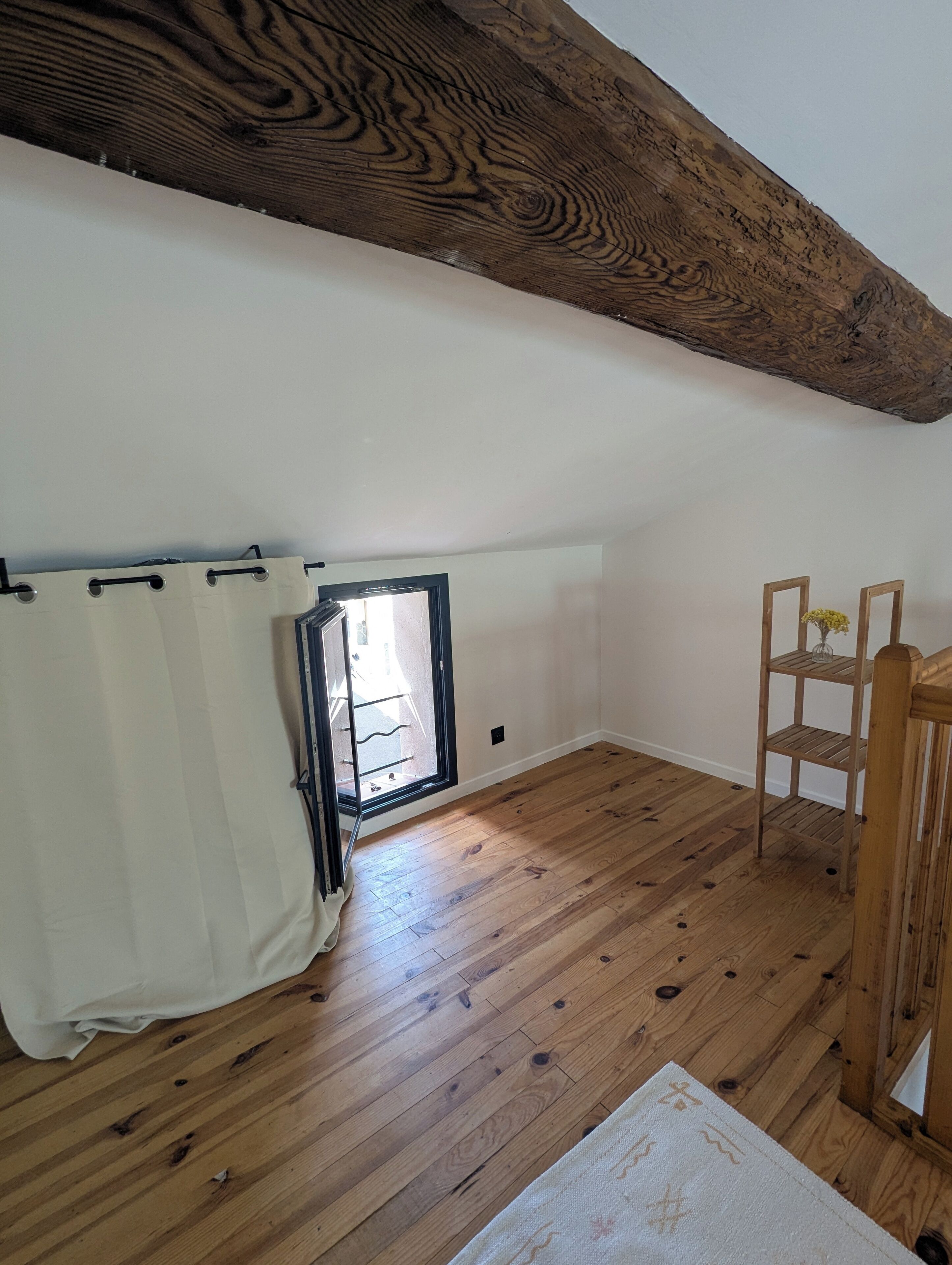 Habitació amb terres de fusta, una finestra amb cortina, una cadira de fusta i una petita estanteria amb flors.