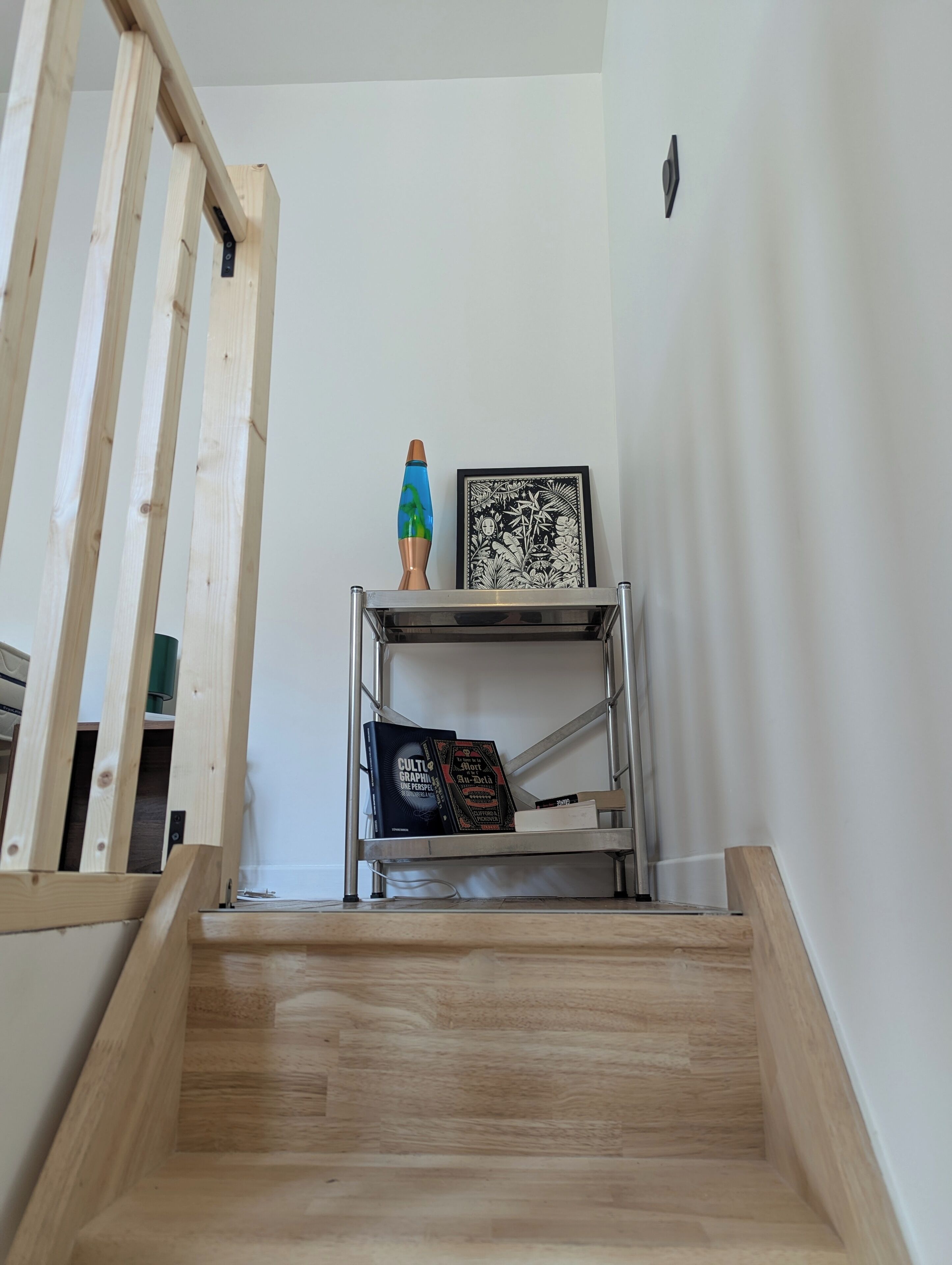 A wooden staircase leading to a room with a metal shelf holding books and a lava lamp.