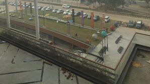A view of a parking lot with several cars, a green space with playground equipment, and a building with a flat roof in the background. - Metro Studio (Greater Noida)