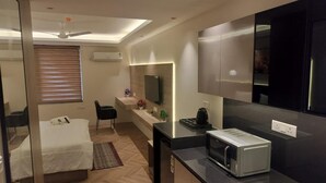 A compact hotel room with a bed, a small kitchenette, a seating area, and a ceiling fan. - Metro Studio (Greater Noida)