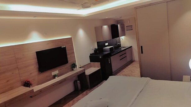 A modern hotel room with a flat-screen TV mounted on a wooden wall, a kitchenette with a black countertop, and a bed with a white mattress. - Metro Studio (Greater Noida)