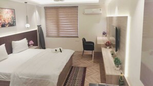 A hotel room with a bed, a desk, a chair, a flat-screen TV mounted on the wall, a ceiling fan, and a window with blinds. - Metro Studio (Greater Noida)