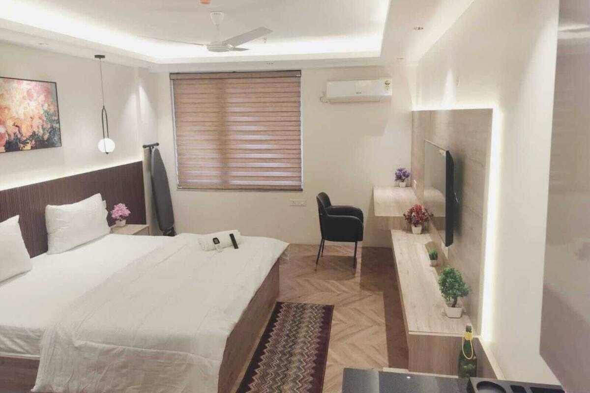 A hotel room with a bed, a desk, a chair, a flat-screen TV mounted on the wall, a ceiling fan, and a window with blinds.