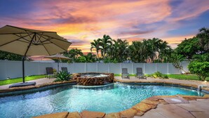 A swimming pool with a hot tub, surrounded by a white fence, and a sunset in the background.