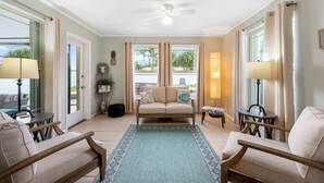 Amplia sala de estar con sofá color beige, dos sillones, una mesa de centro y un ventilador de techo. Grandes ventanas con cortinas dejan entrar luz natural y ofrecen vistas al exterior.
