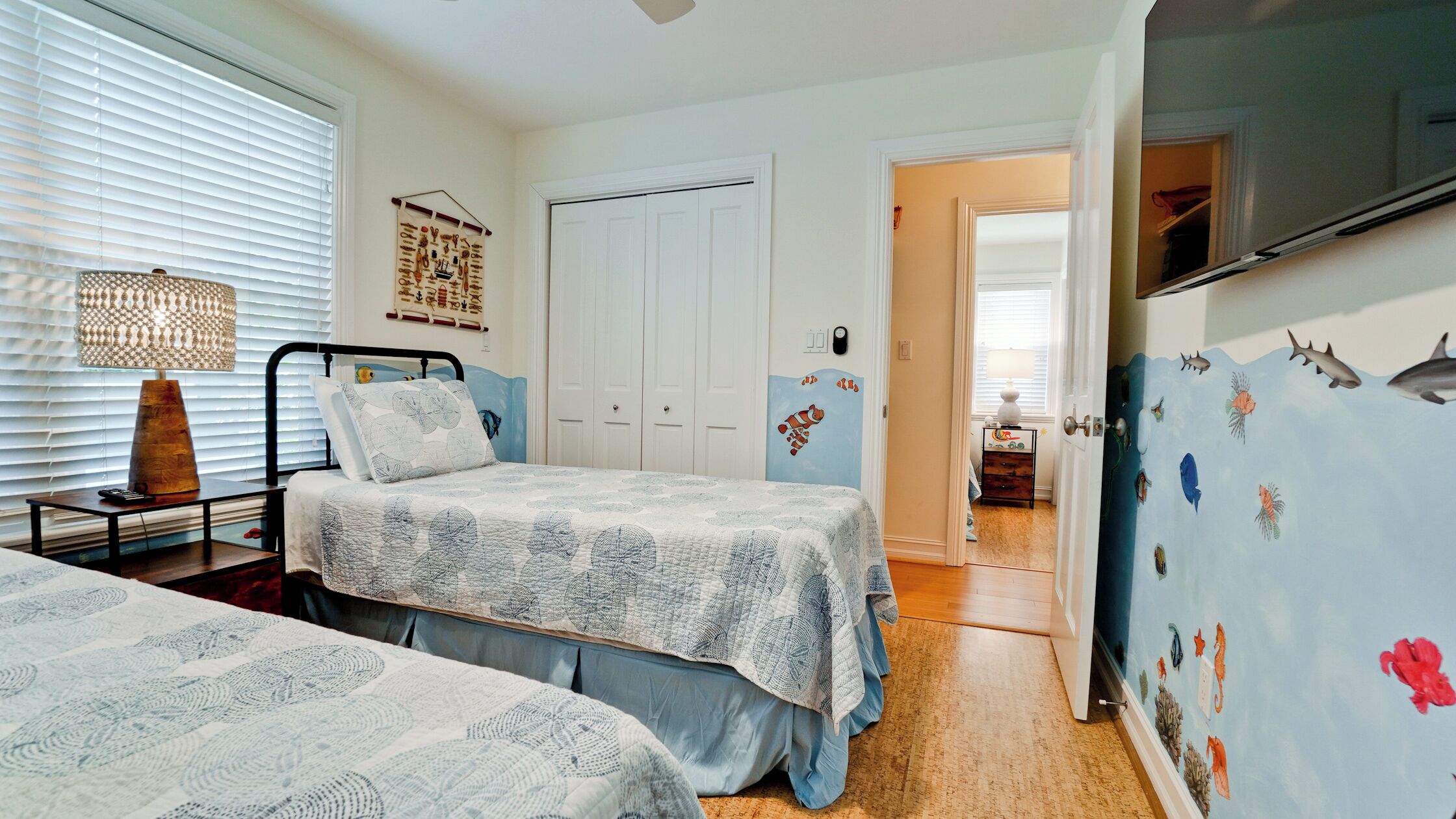 A bedroom with two beds, a bedside table with a lamp, a wall with marine life decals, and a door leading to another room.