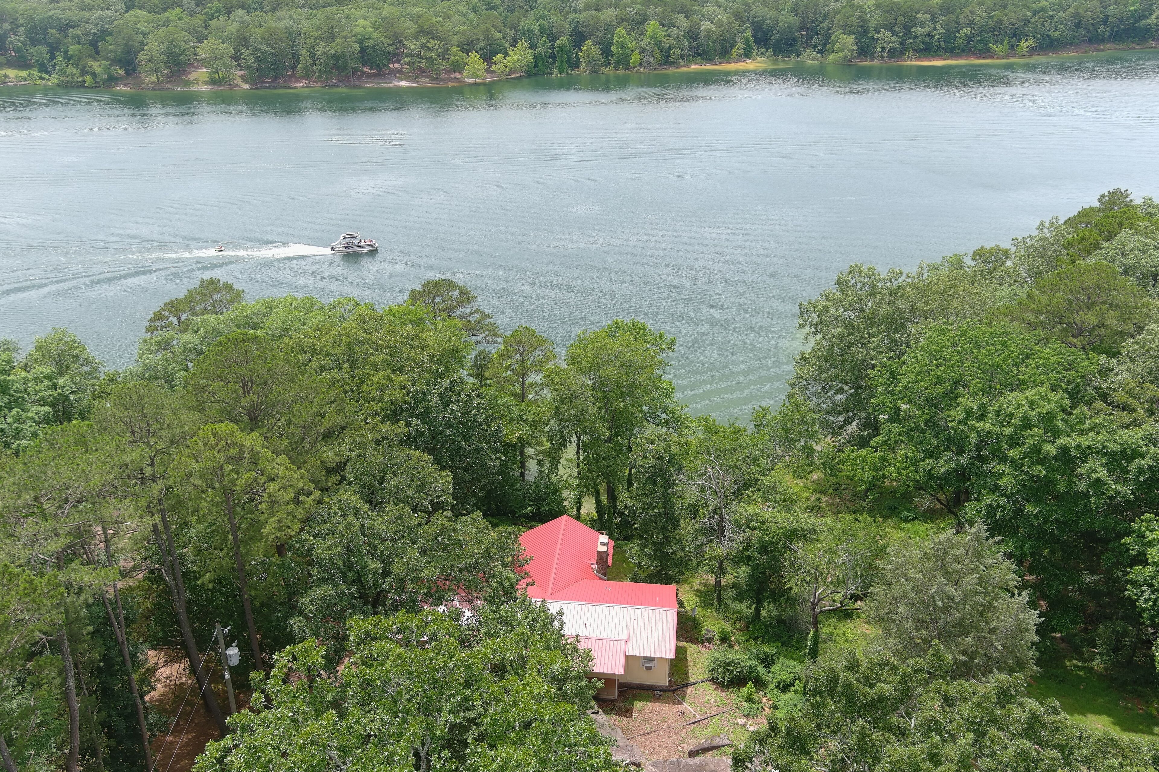A house with a red roof surrounded by dense green trees, overlooking a calm lake with a boat.