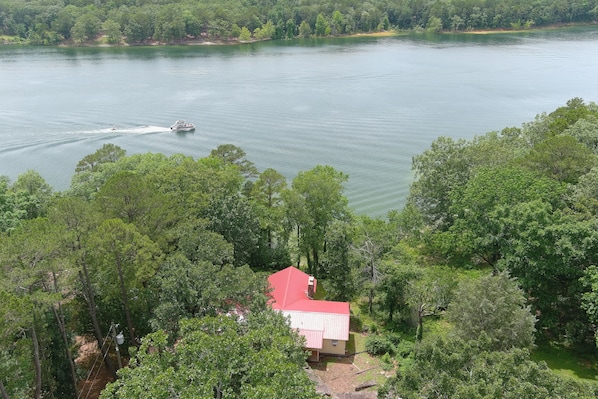 A house with a red roof surrounded by dense green trees, overlooking a calm lake with a boat.