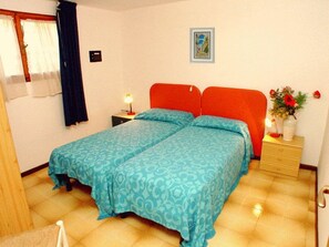 Uma cama de casal com colcha azul estampada, uma mesinha de cabeceira com abajur, um quadro na parede e um vaso com flores na mesinha.