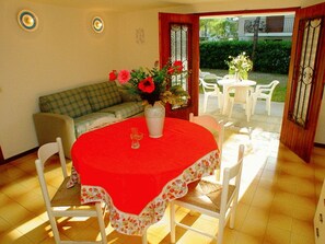 Een eethoek met een rode tafelkleed, witte stoelen, een tafelrunner met bloemmotieven en een vaas met bloemen.