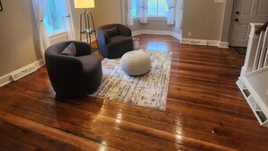 Amplia sala de estar con piso de madera, dos sillones oscuros, un otoman redondo, una lámpara de pie y ventanas grandes con cortinas blancas.
