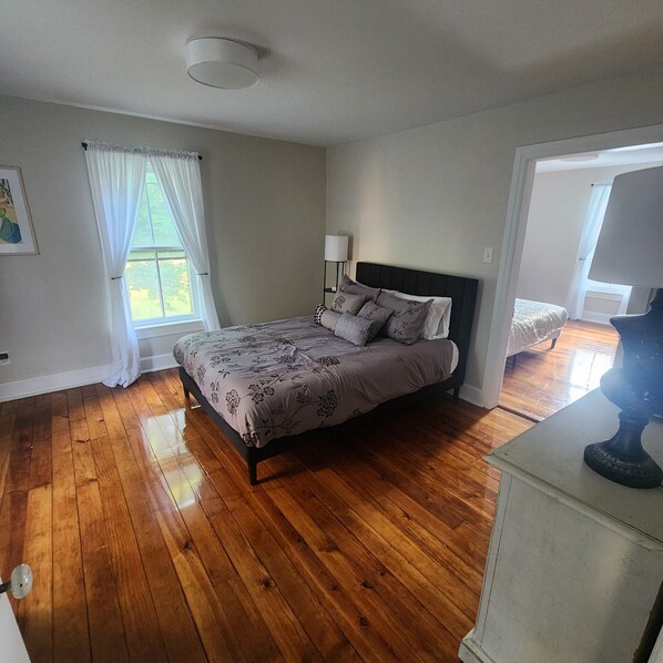 Une chambre avec un plancher en bois, un lit recouvert d’une couvre-lit à motifs floraux, une lampe de chevet et une fenêtre ornée de rideaux légers.