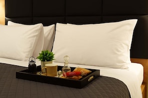 Un lit bien fait, avec des oreillers blancs et une tête de lit noire, un plateau contenant une théière, une tasse, une plante et des pâtisseries.
