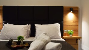 A modern hotel room with a large bed, two white pillows, a black headboard, a bedside table with a lamp, a tray with toiletries, and a small plant. - Lavish Living Suite Heraklion Crete (Hersonissos)