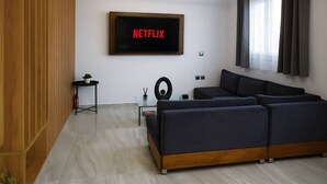 A modern living room with a sectional sofa, a flat-screen TV mounted on the wall, a small side table with a plant, and a window with curtains. - Lavish Living Suite Heraklion Crete (Hersonissos)