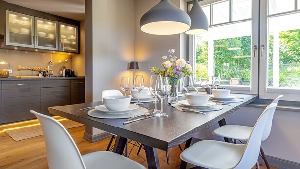 A modern dining area with a long table set for six, white chairs, and a view of greenery through large windows.