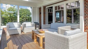 A covered outdoor seating area with wicker furniture, a wooden table, and large windows offering views of greenery.