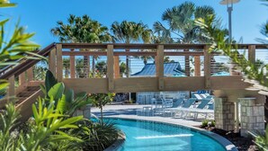 Une piscine d’un bleu éclatant, avec un pont en bois, entourée de palmiers et de chaises longues.