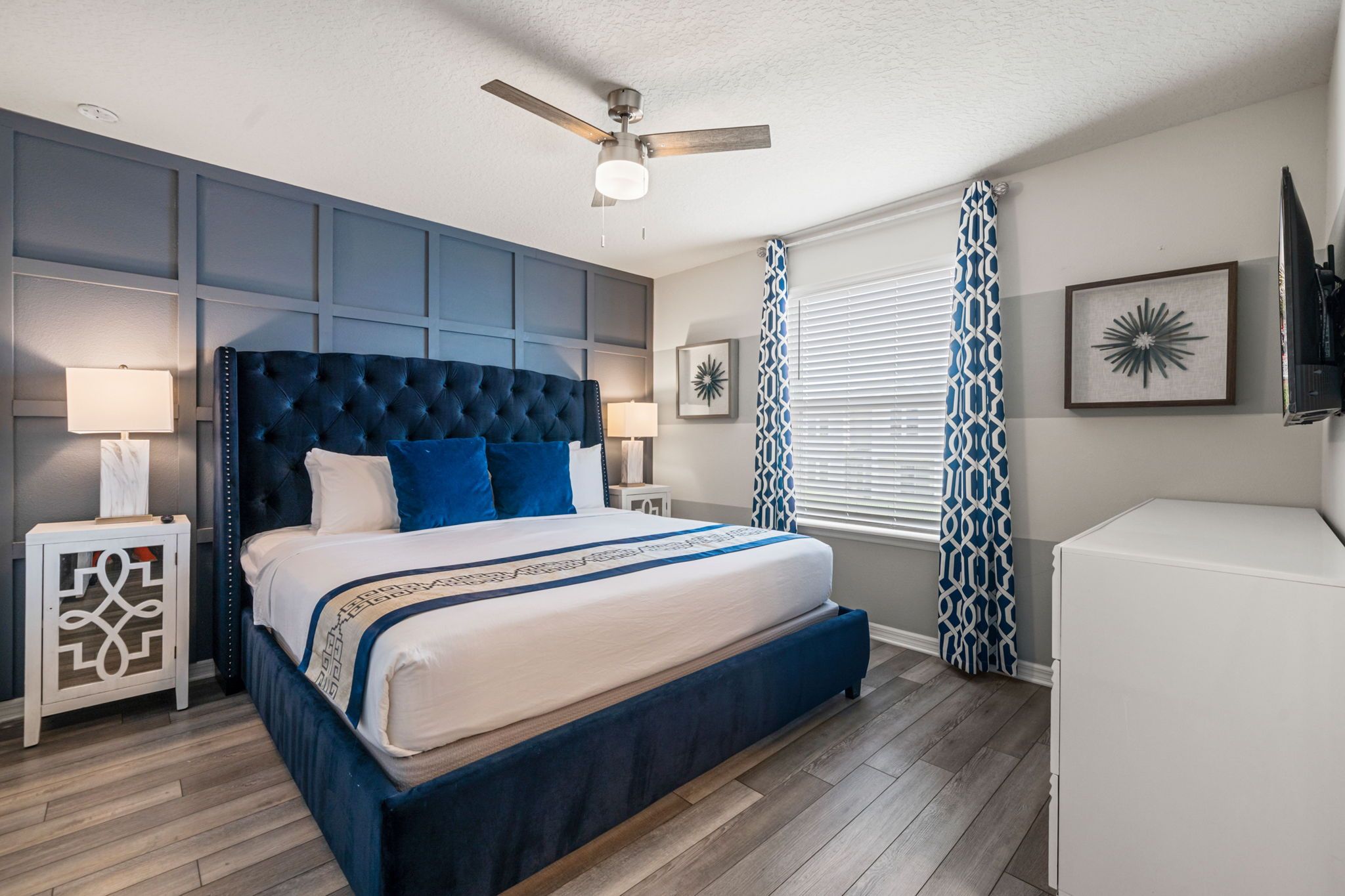A bedroom with a tufted headboard, a large bed, a ceiling fan, a window with patterned curtains, and a flat-screen TV mounted on the wall.