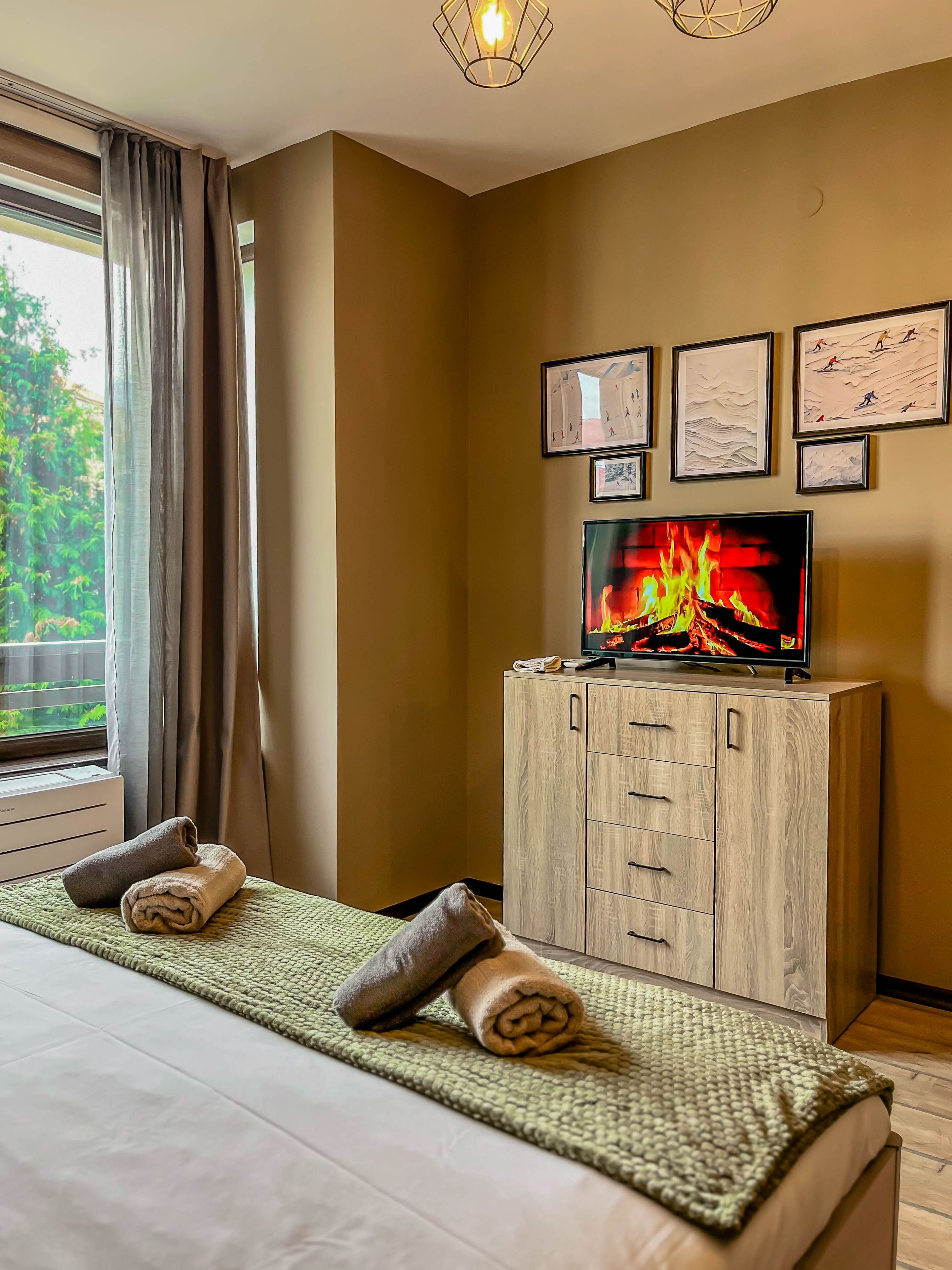 A hotel room with a bed, a television displaying a fire, a wooden cabinet, and a window with curtains.