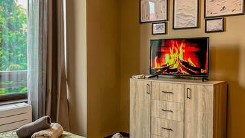 A hotel room with a bed, a television displaying a fire, a wooden cabinet, and a window with curtains.