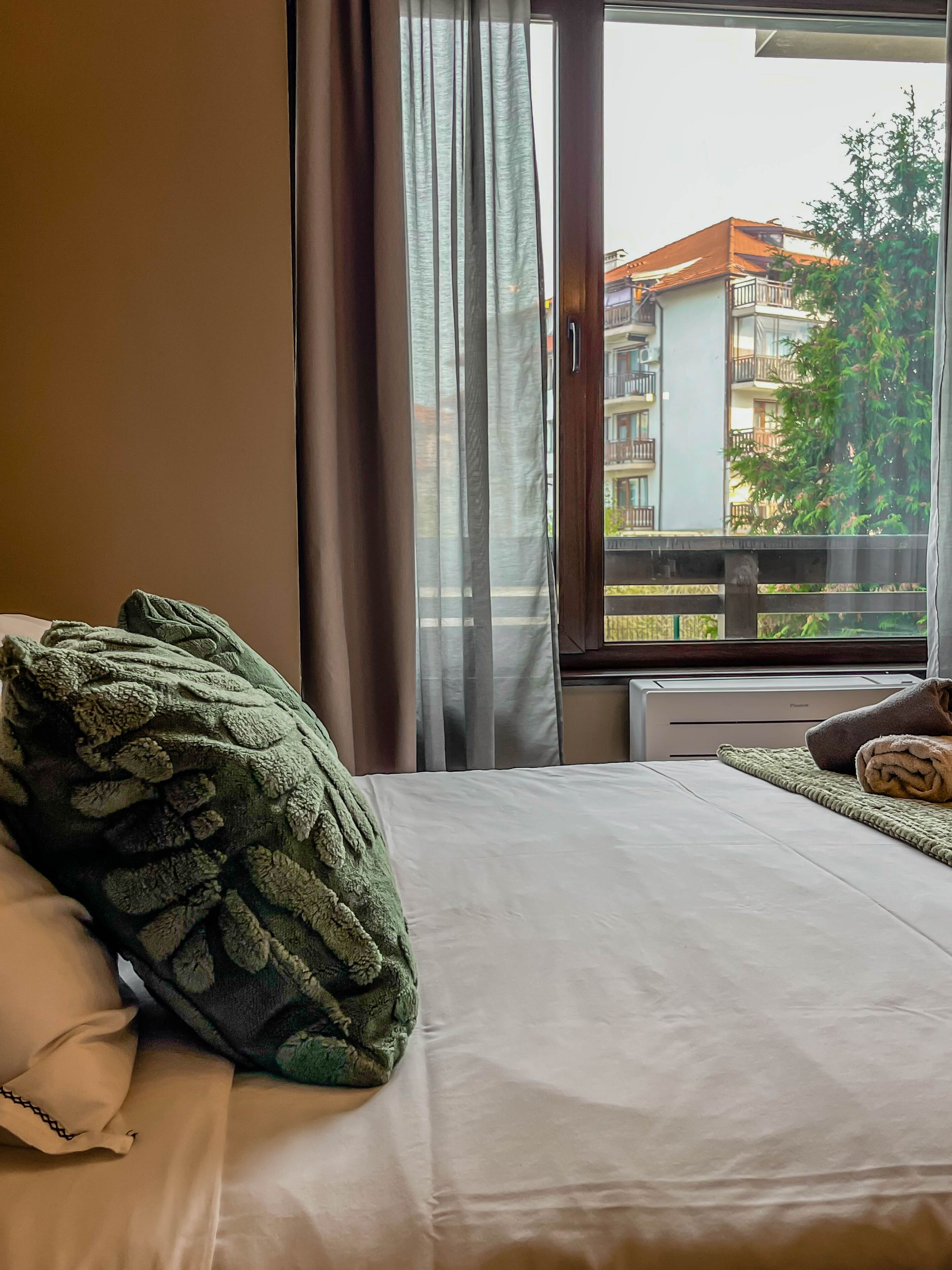 A hotel room with a bed, a window with curtains, and a view of buildings outside.
