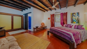 A spacious bedroom with a large bed, wooden ceiling beams, a ceiling fan, and a painting on the wall.