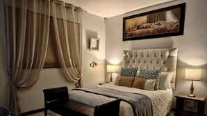Une chambre à coucher avec une tête de lit capitonnée, un lit agrémenté d’oreillers à motifs, une lampe de chevet, un banc et un tableau encadré au mur.